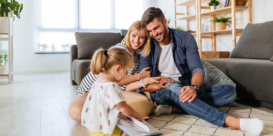 Mutter und Vater sitzen auf dem Teppich und lächeln ihr spielendes Kind an (Foto)